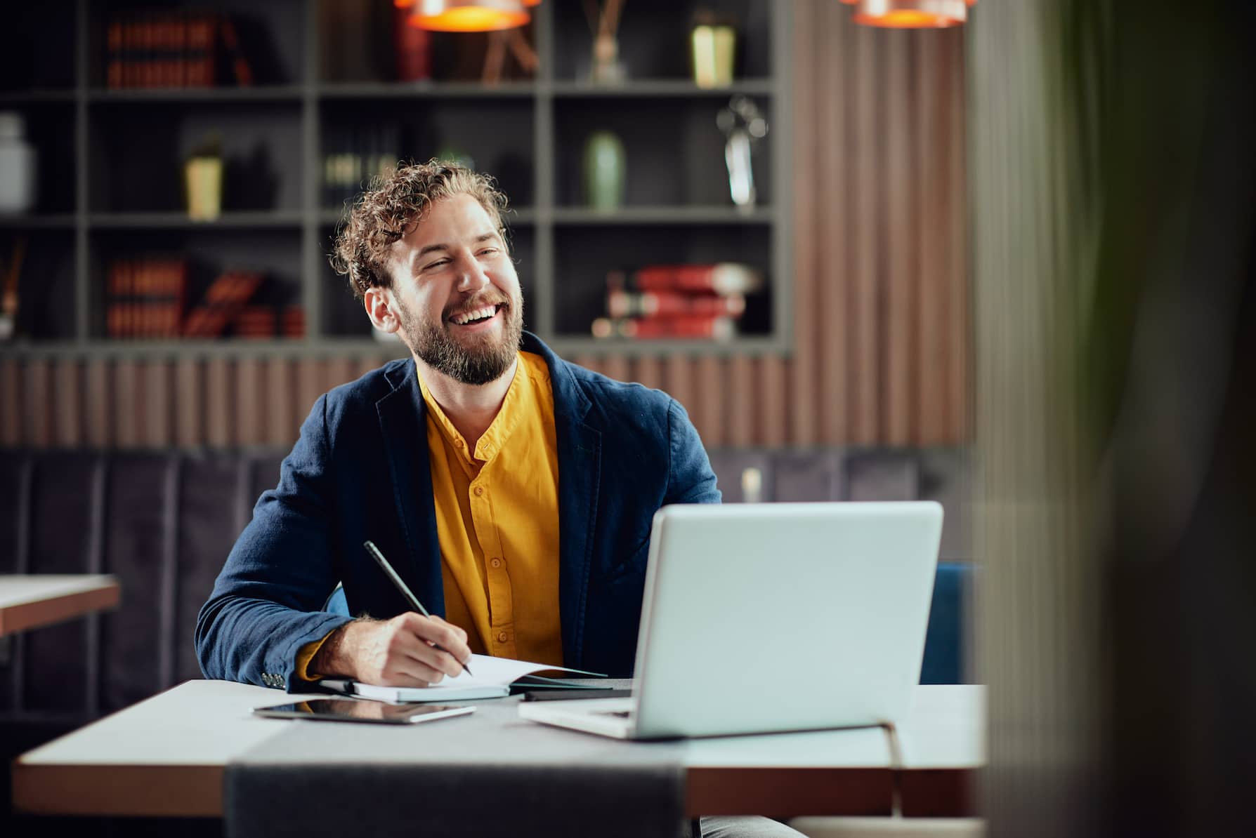 Young smiling bearded man dressed casual writing notes in agenda and looking at laptop while sitting in a table.